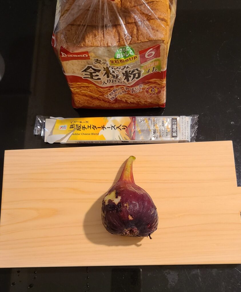 まな板の上に載っている一個の生のいちじくと黒いテーブルの上のベビーチーズと食パン一袋を食材として紹介している様子