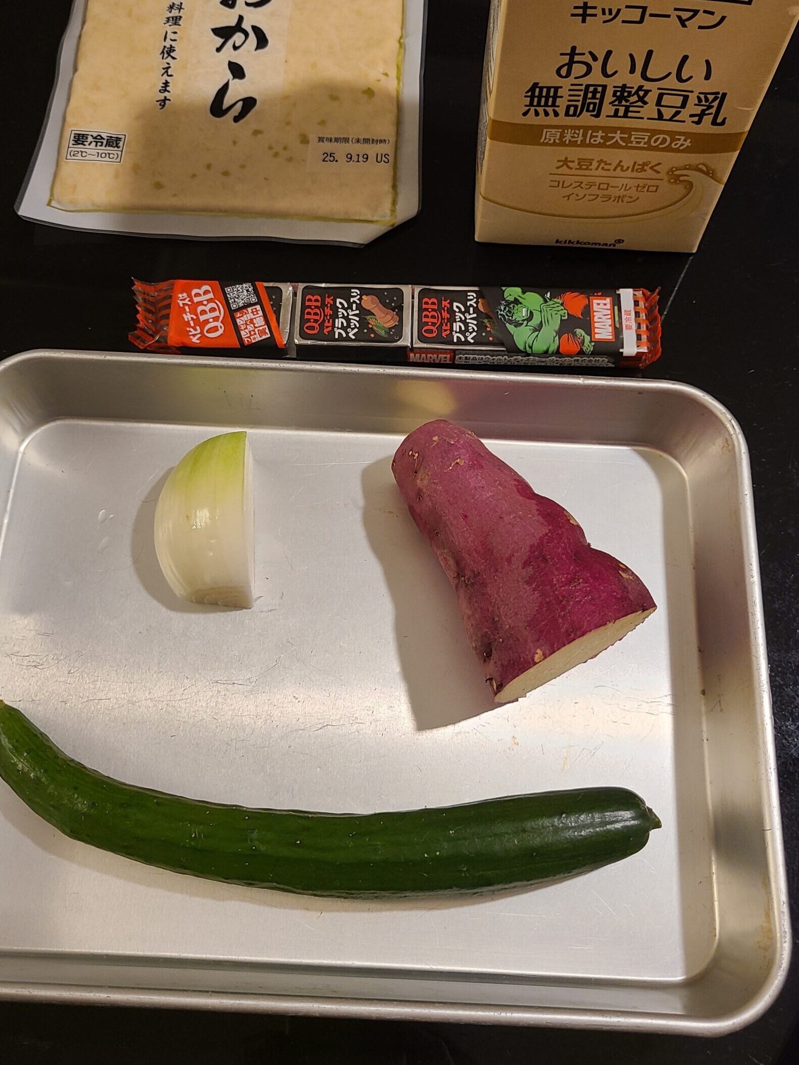 バットに入った1/4の玉ねぎ、1/2のさつま芋、一本のきゅうりとブラックペーパー入りベビーチーズ、生おから、無調整豆乳が黒いテーブルの上にある写真。