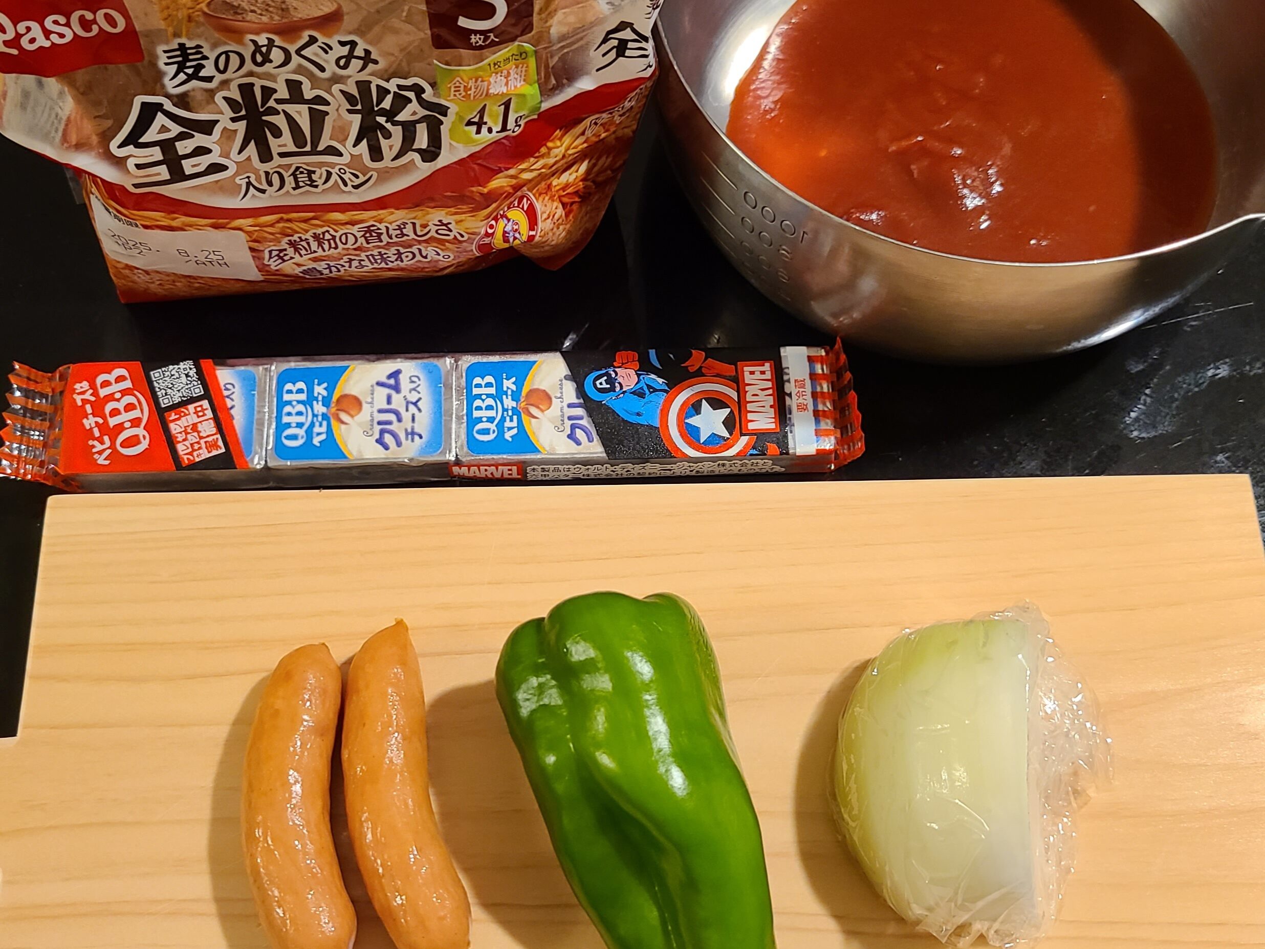 黒いテーブルの上のPascoの麦のめぐみ全粒粉入れ食パン、鍋に入ったトマトソース、QBBベビーチーズクリームチーズ入りと、カッティングボードの上の玉ねぎ1/4、ピーマン1個、ウィンナー2本の写真。