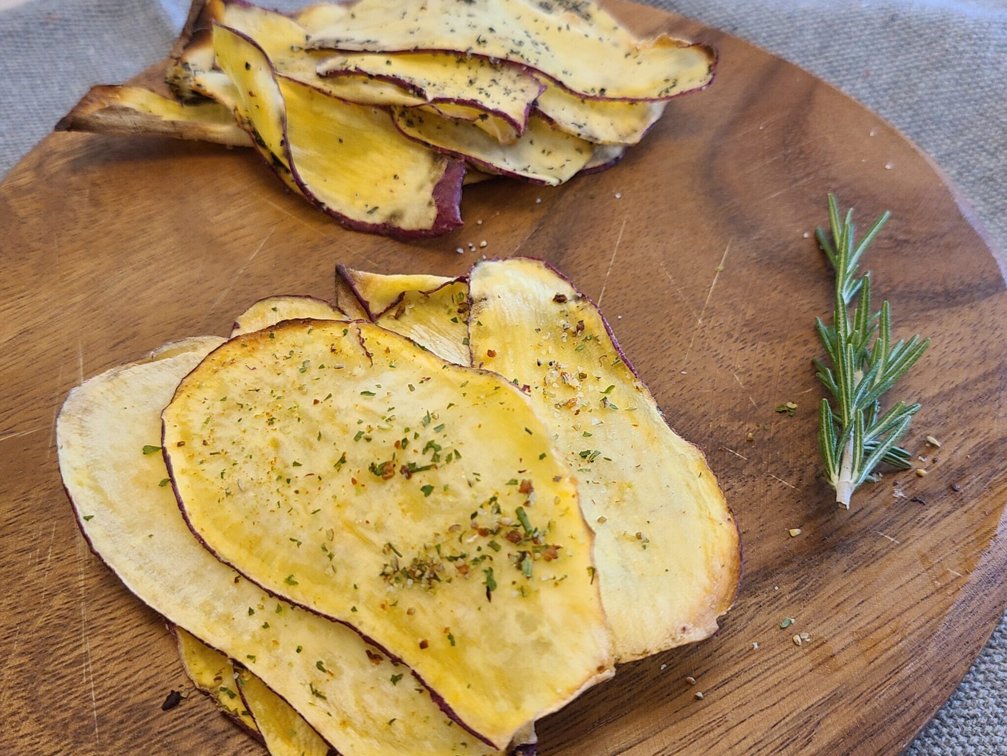 茶色いカッティングボードの上にマジックソルトをかけたさつまいもチップスと、ラカントと黒ごまをかけたさつまいもチップスと、ボードの右端にローズマリーがある写真