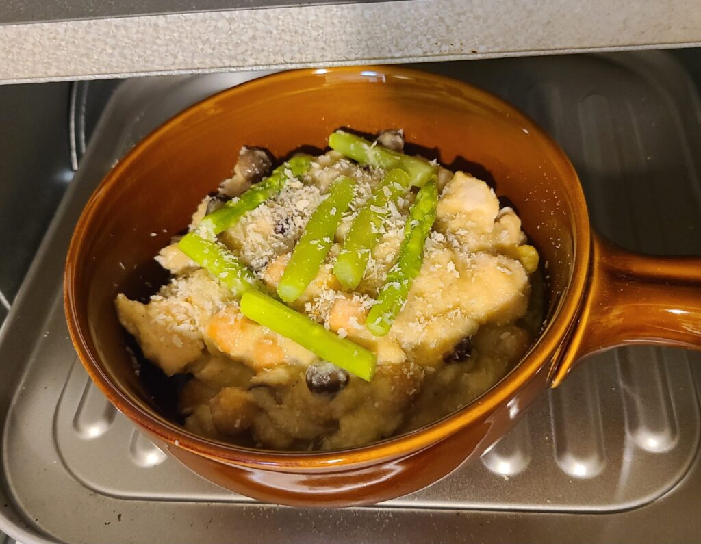 煮込んだ材料をグラタン皿に入れ、アスパラとパン粉をトッピングしトースターに入れている様子